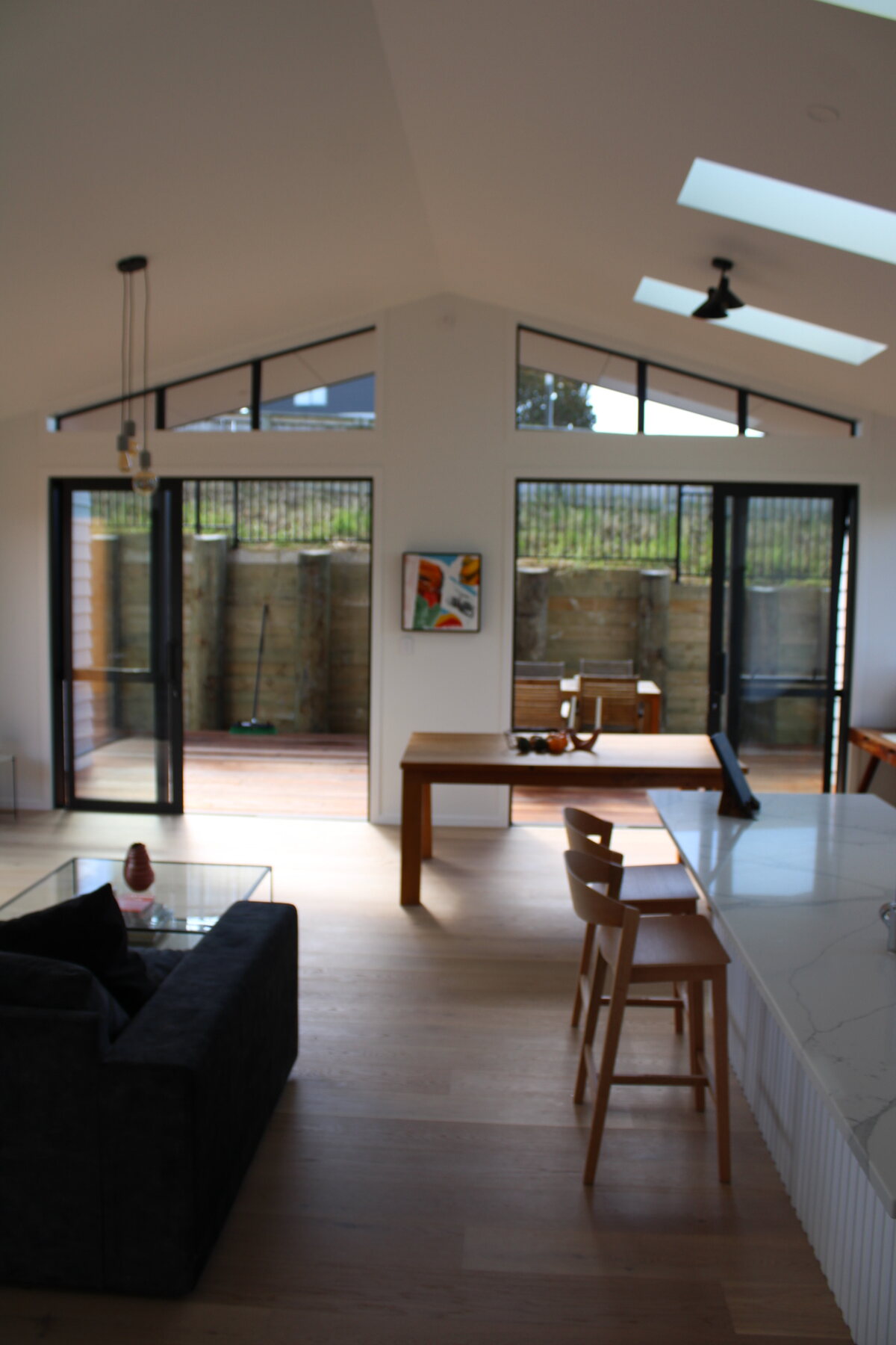 Light open-plan living room with lounge, dining table, kitchen island, skylights, and deck doors